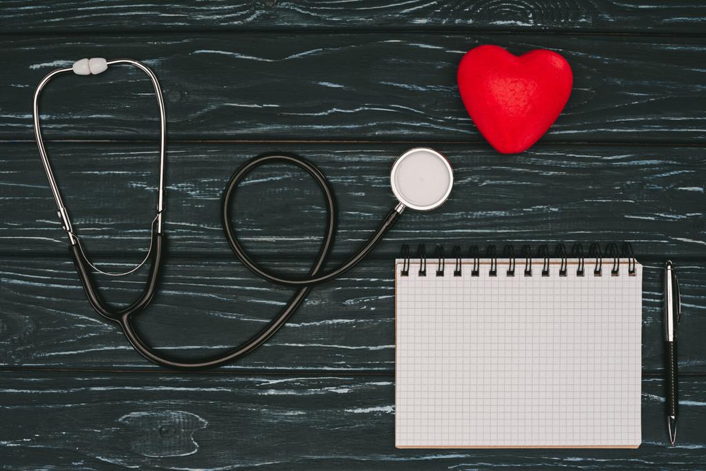 flat lay with arranged red heart, stethoscope and empty notebook on dark wooden tabletop, world health day concept - Photo, Image