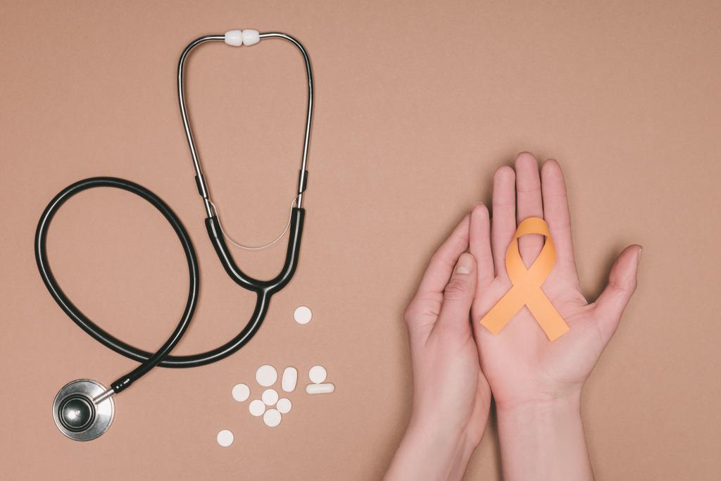 cropped shot of female hands with leukemia, kidney cancer, multiple sclerosis RSD awareness ribbon, pills and stethoscope isolated on beige tabletop - Photo, Image