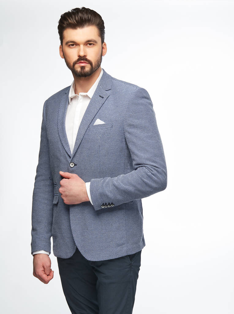 Модный портрет красивого мужчины-модели с темными волосами, бородой и глазами, в белой рубашке, светло-голубой куртке и темных брюках, позируя на белом фоне
 - Фото, изображение