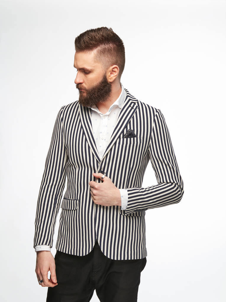 Fashion portrait of handsome male model with dark hair, beard and eyes, wearing white shirt, black and white striped jacket and dark pants, posing on white background - Photo, Image