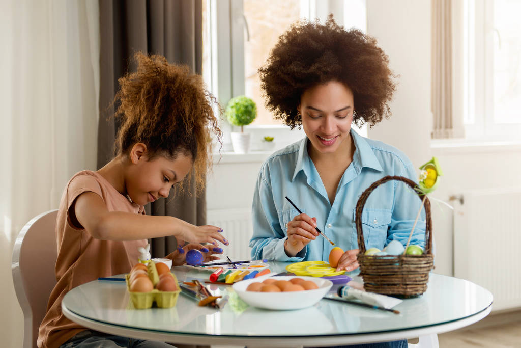美しいアフリカ系アメリカ人の女性と彼女の娘がテーブルでイースターの卵を着色   - 写真・画像