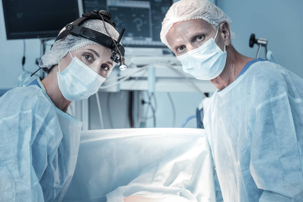 Médicos responsáveis qualificados olhando em linha reta e de pé perto da mesa de operação
. - Foto, Imagem