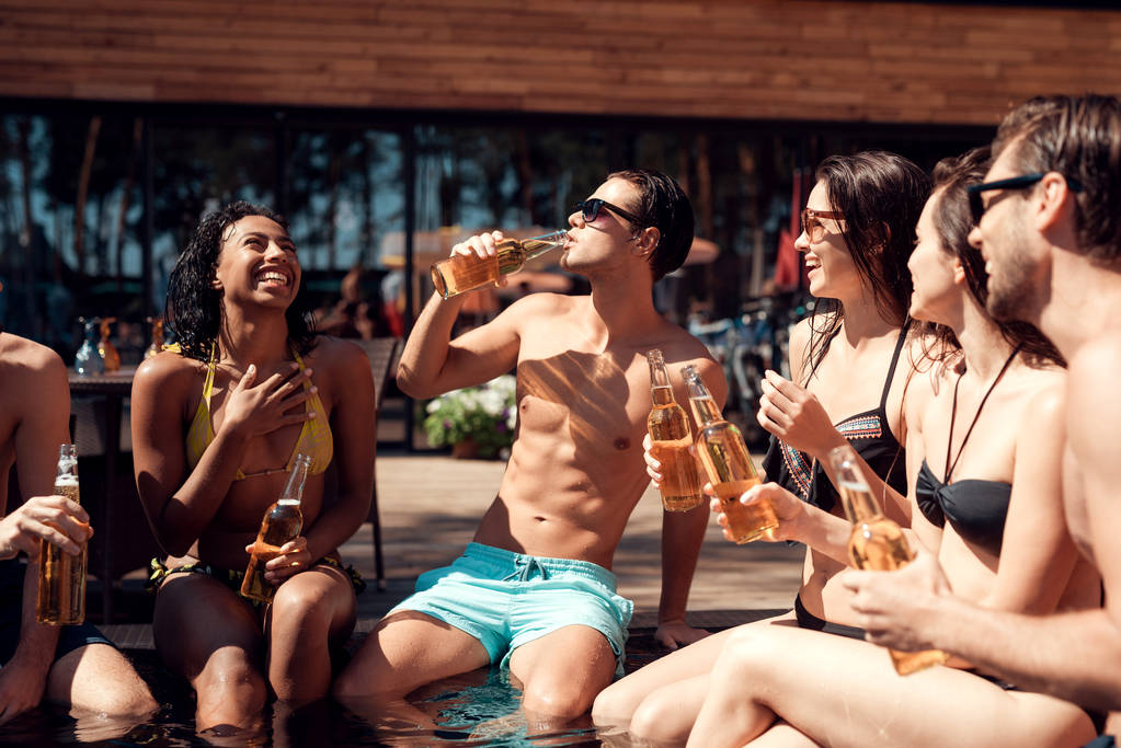 Jóvenes amigos animando con botellas de cerveza en la piscina en verano. Fiesta en la piscina. Empresa multiétnica de amigos en piscina
. - Foto, Imagen
