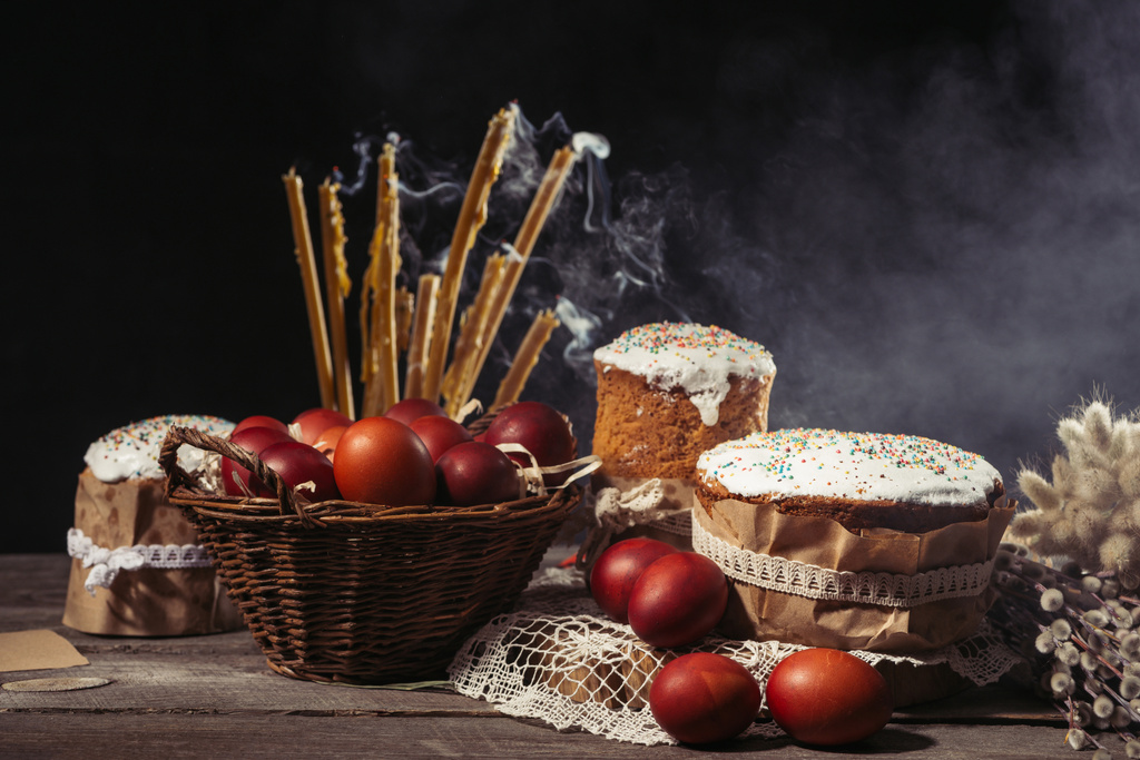 painted eggs in basket, candles with smoke and homemade easter cakes on wooden table - Photo, Image