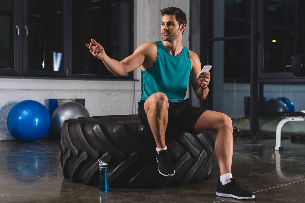 Αθλητικός τύπος κάθεται στο λάστιχο και χρησιμοποιώντας το smartphone στο γυμναστήριο - Φωτογραφία, εικόνα
