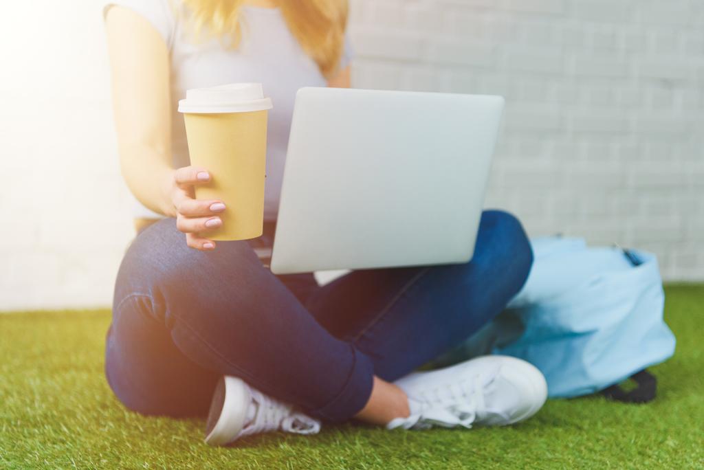 recortado disparo de mujer joven sentado en la hierba con taza de café para ir y trabajar con el ordenador portátil
 - Foto, Imagen