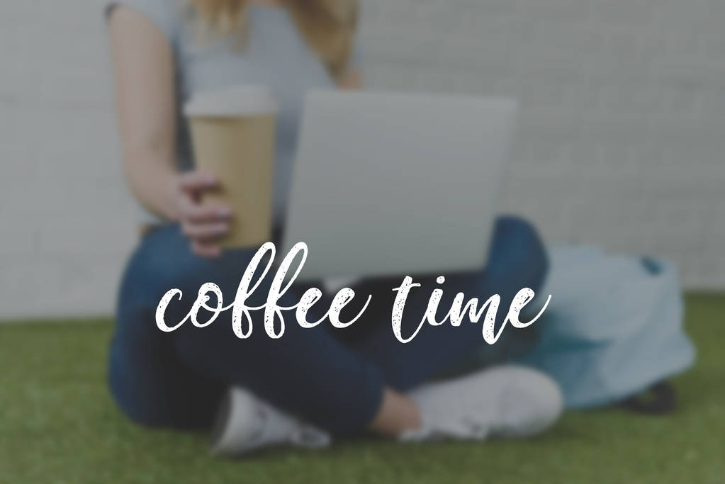 tiro cortado de mulher jovem sentado na grama com xícara de café para ir e trabalhar com laptop, inscrição tempo café
 - Foto, Imagem