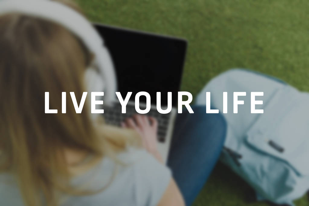 high angle view of young woman sitting on grass with headphones on head and working with laptop, live your life inscription - Photo, Image