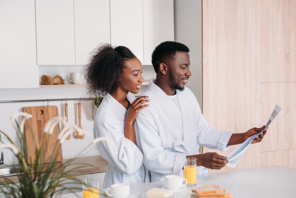 Glimlachen van Afro-Amerikaanse man lezing krant terwijl vriendin hem omhelzen in keuken - Foto, afbeelding