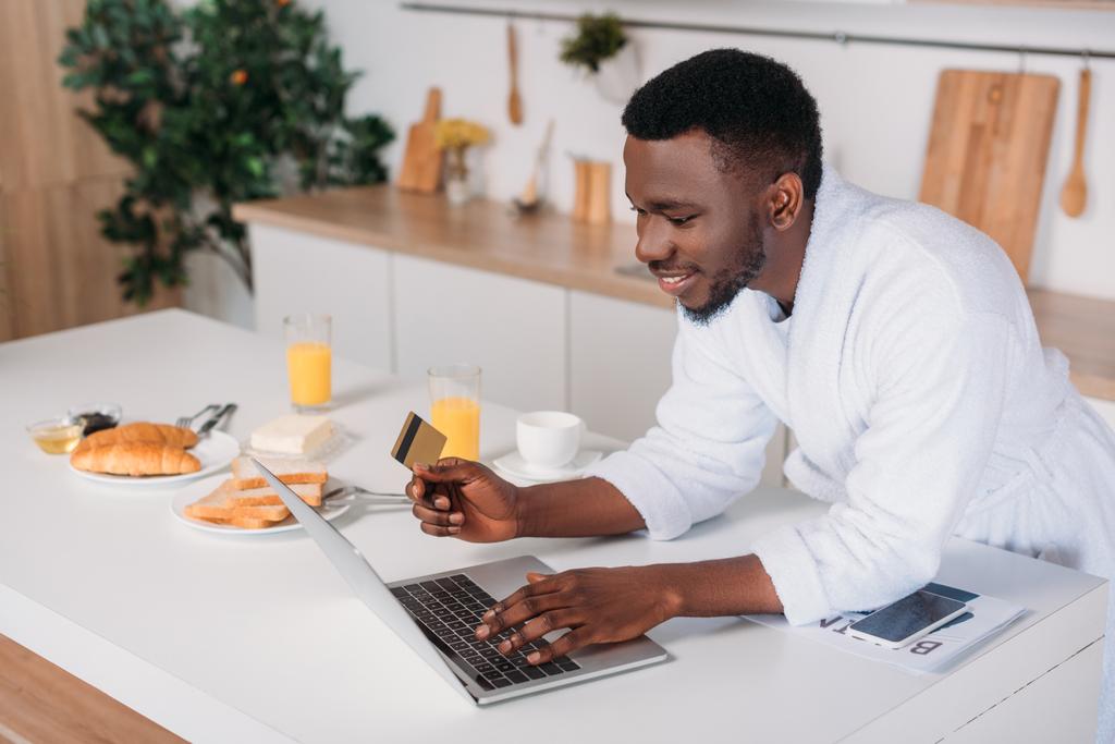 African american man doing online shopping and standing with credit card in kitchen - Photo, Image