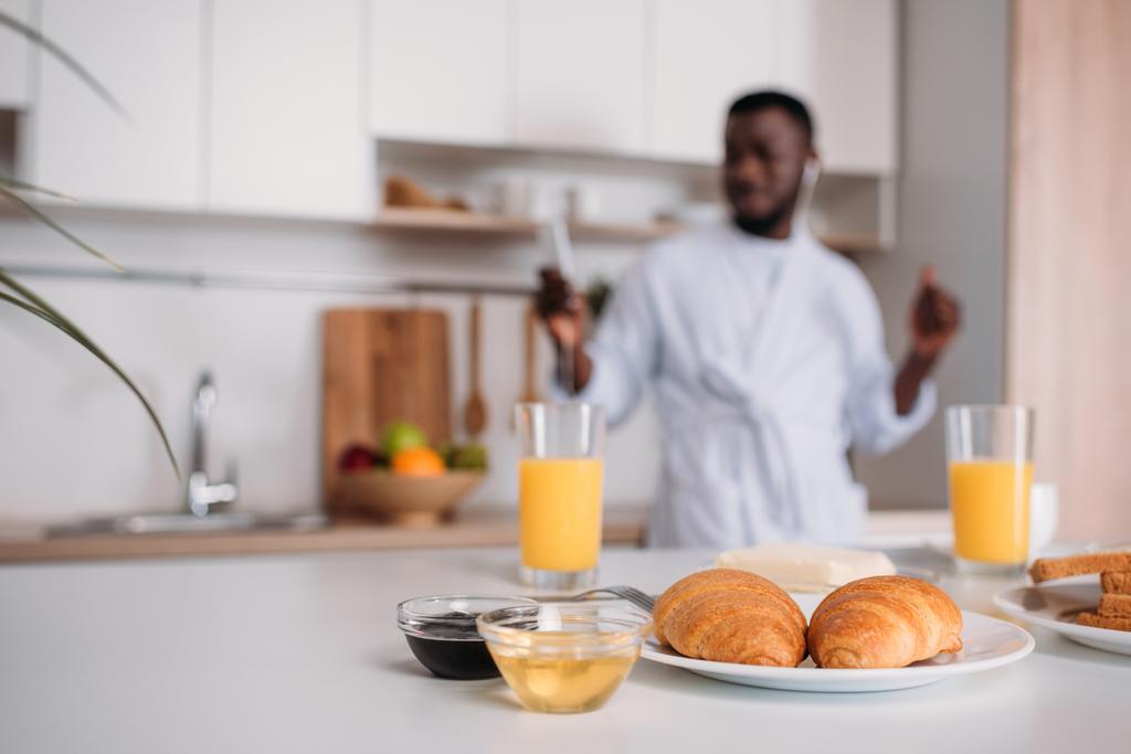 Close up view of croissants on plate, orange juice, jams and butter with young man in earphones on blurred background - Photo, Image