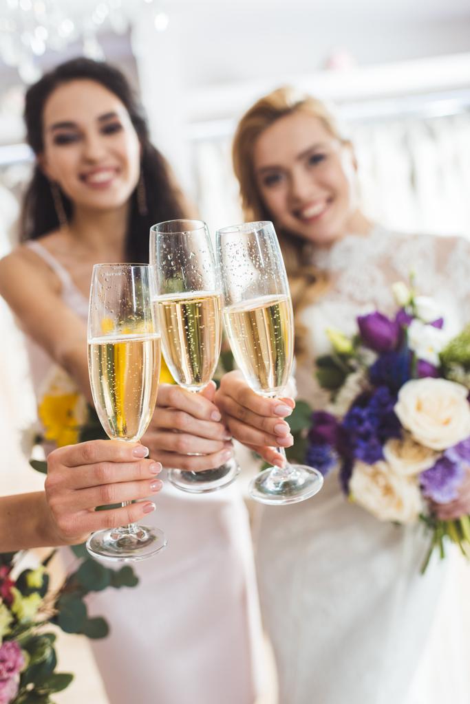 Молодые улыбающиеся невесты с шампанским в очках в свадебном ателье
 - Фото, изображение