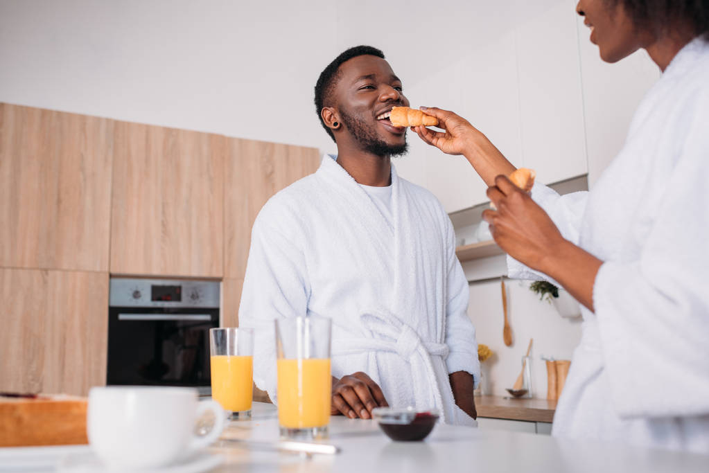 Low angle view of african american woman feeding boyfriend by croissant in kitchen - Photo, Image