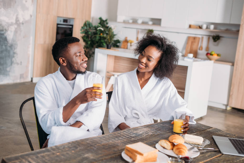Lachende paar zittend aan tafel met ontbijt en laptop in de keuken - Foto, afbeelding