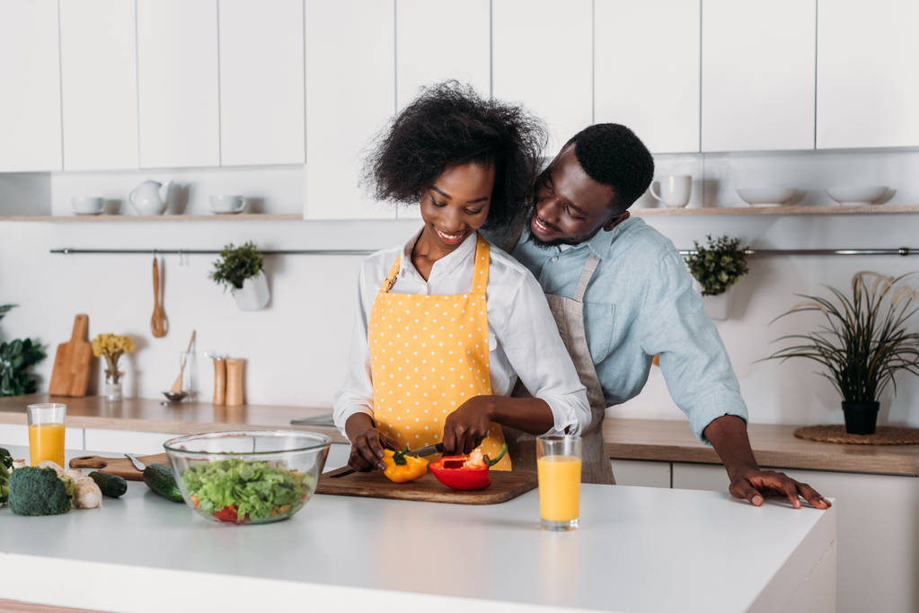 Усміхнений чоловік обіймає дівчину, коли вона ріже перець на дошці на кухні
 - Фото, зображення