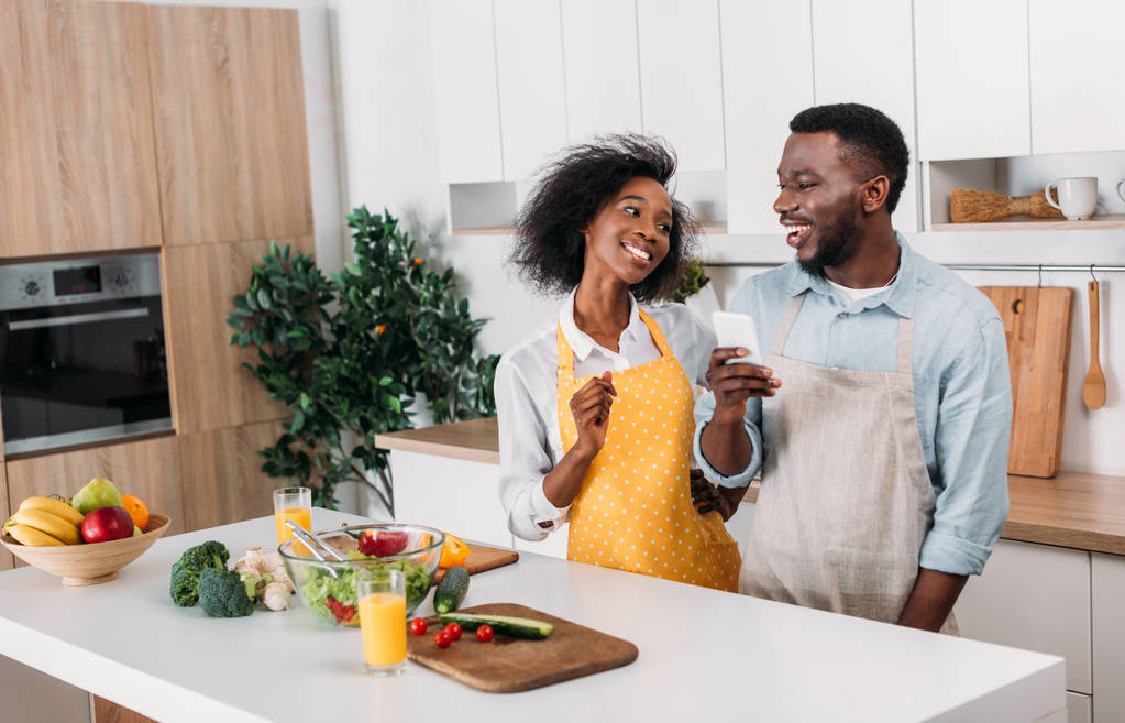 Afrikkalainen amerikkalainen pari esiliinat seisoo pöydässä ruoan kanssa ja käyttää älypuhelinta keittiössä
 - Valokuva, kuva