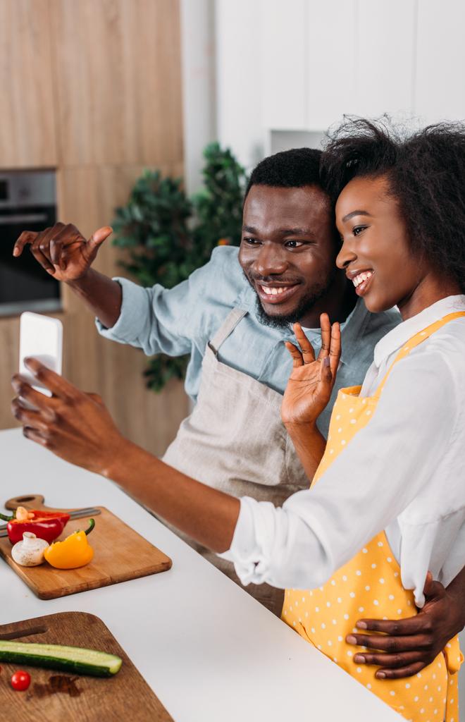Jeune couple afro-américain en tabliers prenant selfie dans la cuisine
 - Photo, image
