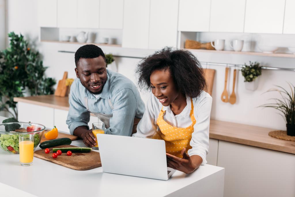 Hymyilevä nuori pari käyttää kannettavaa tietokonetta pöydässä vihannesten kanssa keittiössä
 - Valokuva, kuva