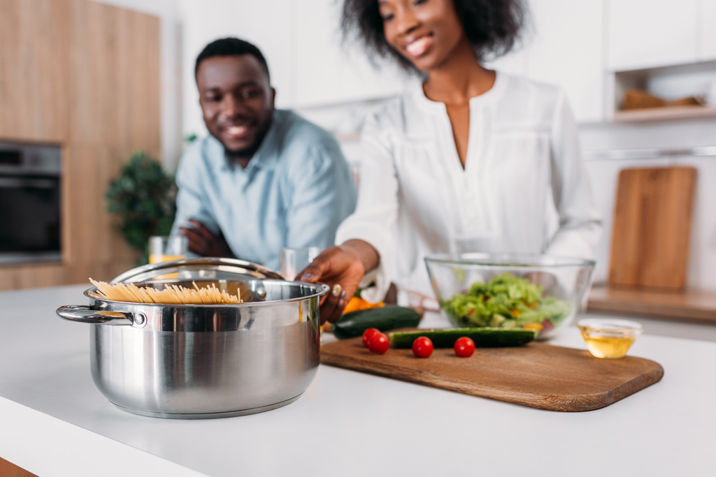 Nahaufnahme des Kochtopfes mit Nudeln und Gemüse am Tisch mit lächelndem Paar dahinter - Foto, Bild
