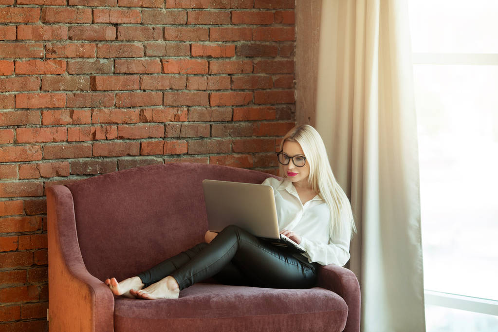 hermosa joven con el pelo rubio sentado en el sofá con una computadora portátil en la mano cerca de una pared de ladrillo
 - Foto, imagen