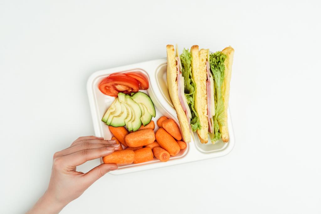 обрезанное изображение женщины, берущей морковь из коробки с обедом, изолированной на белом
 - Фото, изображение