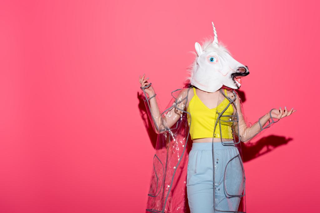 grappige vrouw in modieuze transparante regenjas en unicorn masker op rood - Foto, afbeelding