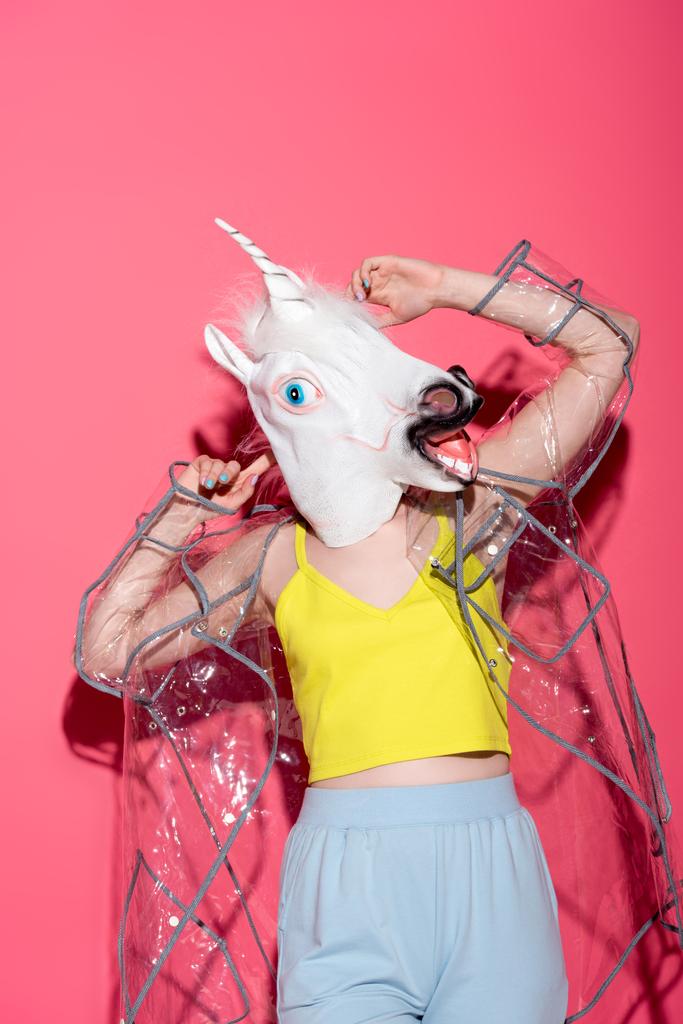 ridiculous woman in fashionable transparent raincoat and unicorn mask on red - Photo, Image