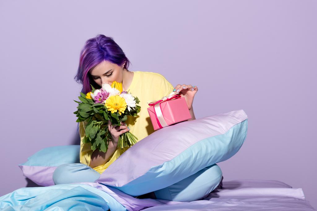 ギフト用の箱とベッドに座って花を持つ美しい若い女性 - 写真・画像