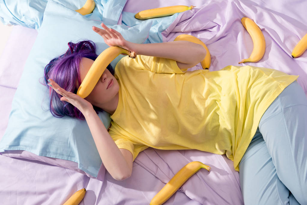 hoge hoekmening van jonge vrouw liggend in bed met bananen en die betrekking hebben op gezicht - Foto, afbeelding