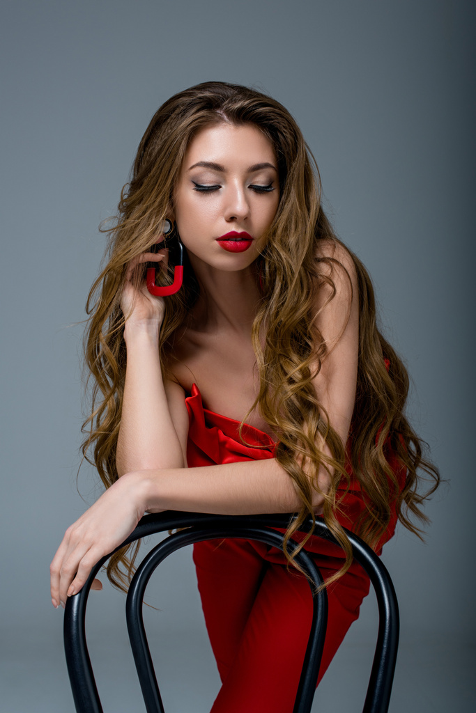 ελκυστική γυναίκα με μακριά μαλλιά ποζάρει στα κόκκινα ρούχα στην καρέκλα, απομονώνονται σε γκρι - Φωτογραφία, εικόνα