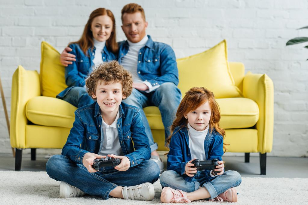 Lapset pelaavat videopeliä, kun vanhemmat istuvat sohvalla
 - Valokuva, kuva
