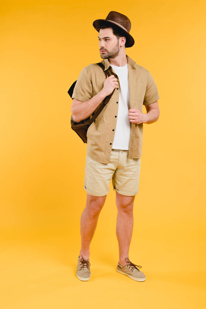 Вид в полный рост красивого молодого человека в шортах и шляпе, держащего рюкзак и уединяющегося на желтом
 - Фото, изображение