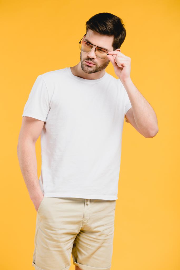 όμορφος γενειοφόρος νέος σε λευκό t-shirt προσαρμογή γυαλιά ηλίου και αναζητούν μακριά απομονώνονται σε κίτρινο - Φωτογραφία, εικόνα