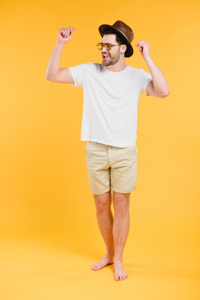веселый молодой босоногий мужчина в шортах и солнцезащитных очках, смотрящий в сторону, изолированный на желтый
 - Фото, изображение