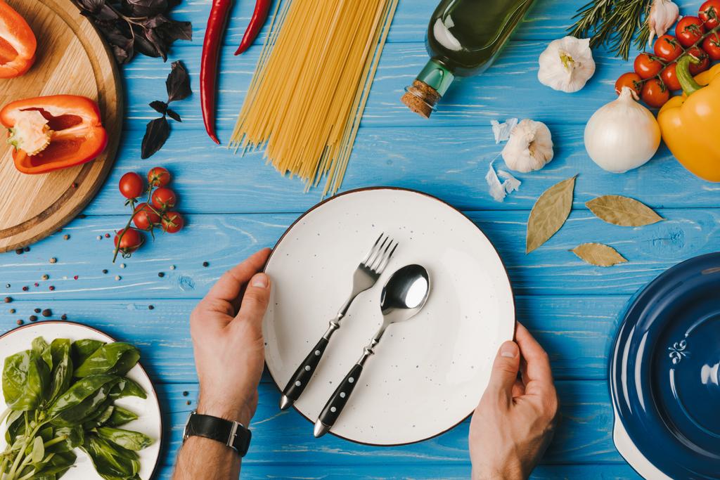 περικομμένη εικόνα ανθρώπου βάζοντας το πιάτο με πιρούνι και κουτάλι σε μπλε πίνακα - Φωτογραφία, εικόνα