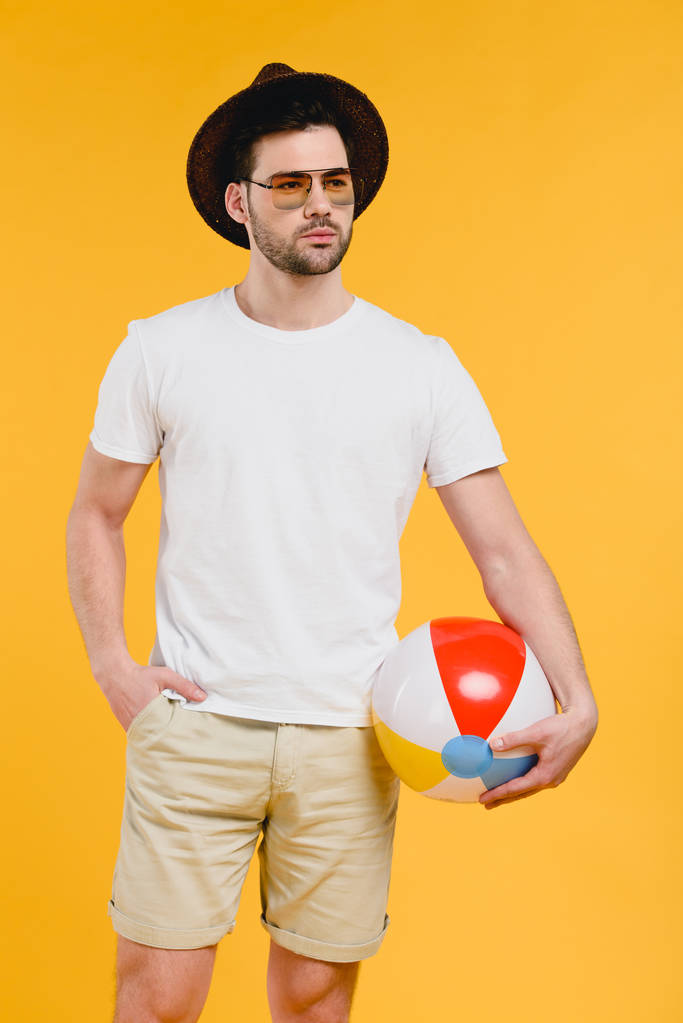 młody człowiek w kapelusz i okulary przeciwsłoneczne, trzymając piłka plażowa i patrząc od hotelu na białym kolorem żółtym - Zdjęcie, obraz