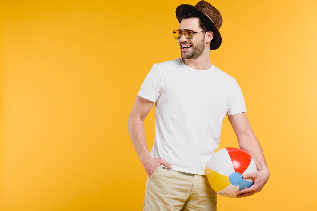 όμορφος χαμογελαστός νεαρός άνδρας στο καπέλο και γυαλιά ηλίου κρατώντας φουσκωτή μπάλα και αναζητούν μακριά απομονώνονται σε κίτρινο - Φωτογραφία, εικόνα