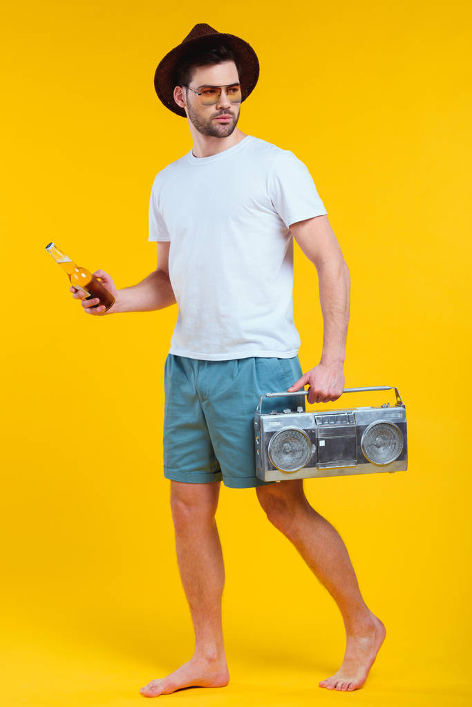 молодой босиком в шортах с магнитофоном и бутылкой летнего напитка, изолированного на желтом
 - Фото, изображение