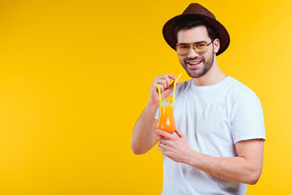 guapo joven en sombrero y gafas de sol bebiendo cóctel de verano y sonriendo a la cámara aislado en amarillo
 - Foto, imagen