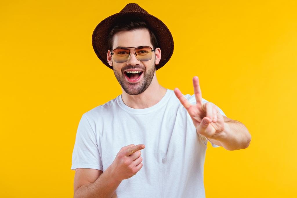 joven alegre en camiseta blanca, sombrero y gafas de sol mostrando signo de victoria y señalando con el dedo aislado en amarillo
 - Foto, imagen