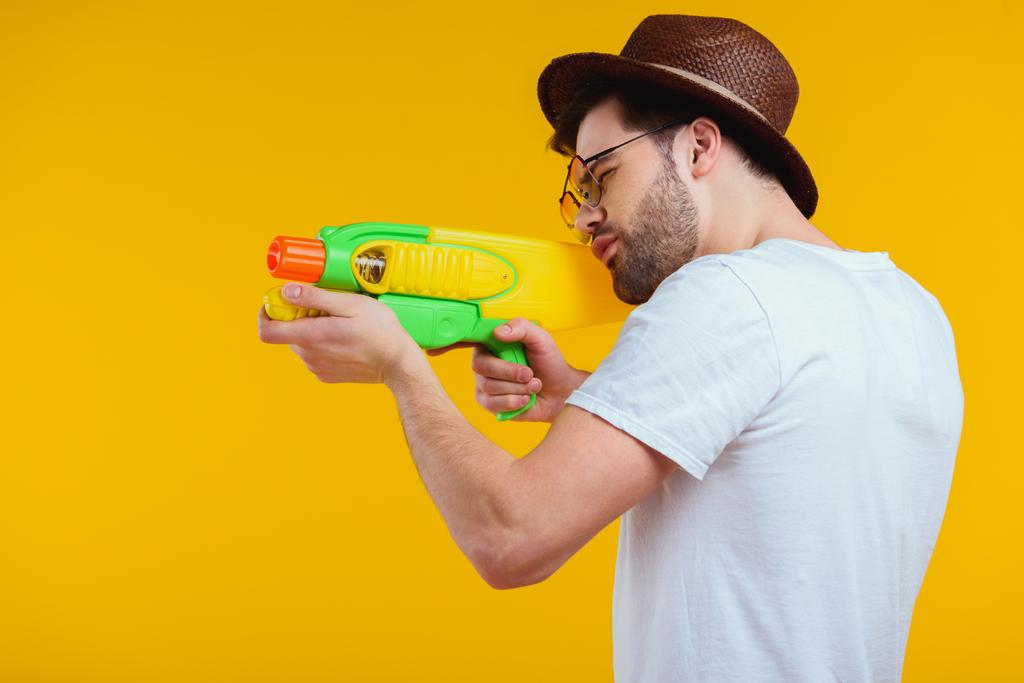 side view of bearded young man in hat and sunglasses shooting with water gun isolated on yellow - Photo, Image