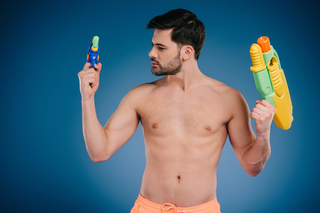 barbuto giovane uomo petto nudo in possesso di pistole ad acqua su blu
 - Foto, immagini