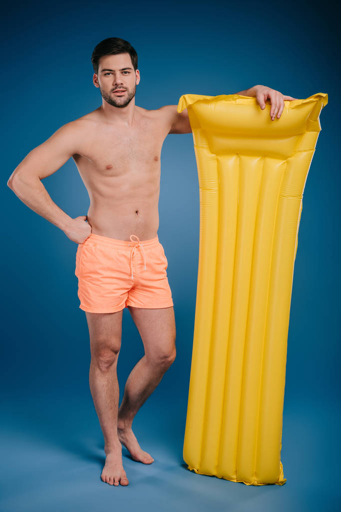 knappe jonge man in korte broek permanent met zwemmen matras en camera op blauw - Foto, afbeelding