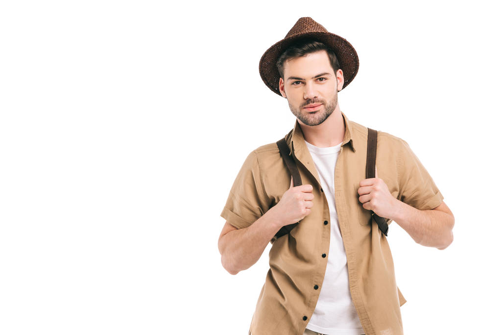 pohledný mladý cestovatel v klobouku drží batoh a při pohledu na fotoaparát izolované na bílém - Fotografie, Obrázek