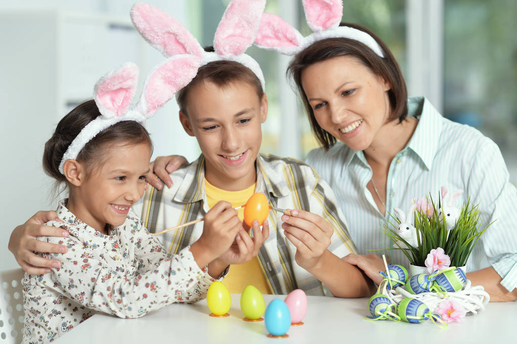 Mãe com seu filho e filha vestindo orelhas de coelho pintando ovos para a Páscoa
 - Foto, Imagem