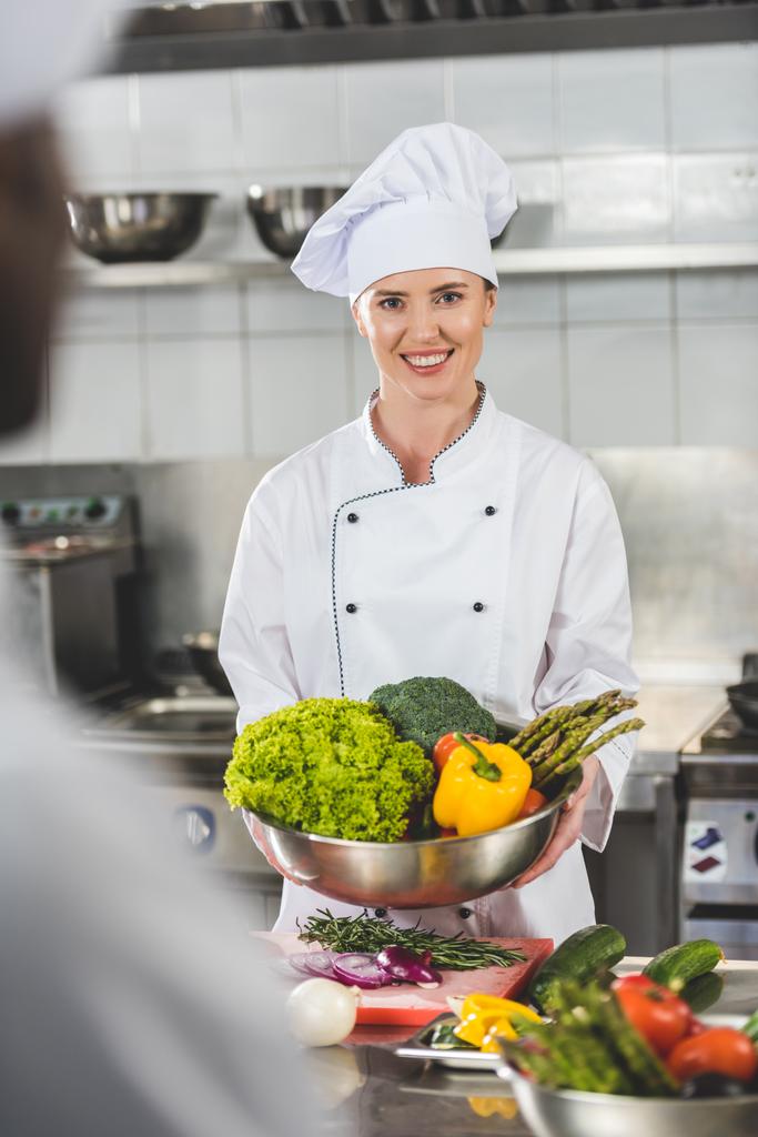 Chef souriant tenant bol avec des légumes et regardant la caméra à la cuisine du restaurant
 - Photo, image