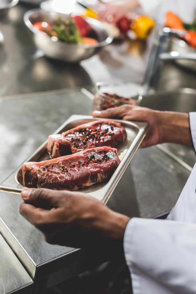 abgeschnittenes Bild eines afrikanisch-amerikanischen Kochs mit Tablett mit rohem Fleisch in der Restaurantküche  - Foto, Bild
