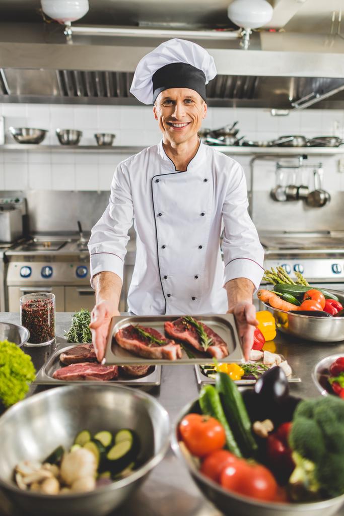 улыбающийся шеф-повар берет поднос с сырым мясом на кухне ресторана
 - Фото, изображение