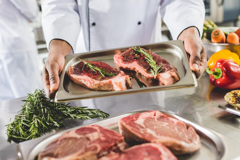 oříznutý obraz afrických amerických šéfkuchaře drží podnos s syrové steaky v restauraci kitchen  - Fotografie, Obrázek