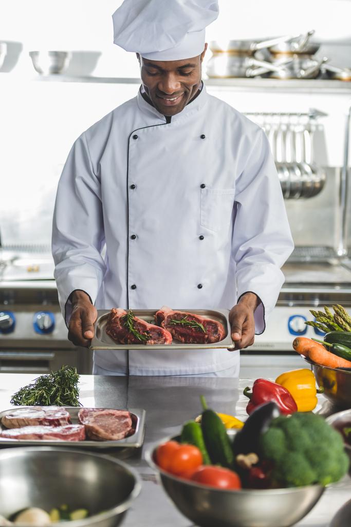 усміхнений красивий афроамериканський шеф-кухар тримає лоток з невареним м'ясом на кухні ресторану
 - Фото, зображення